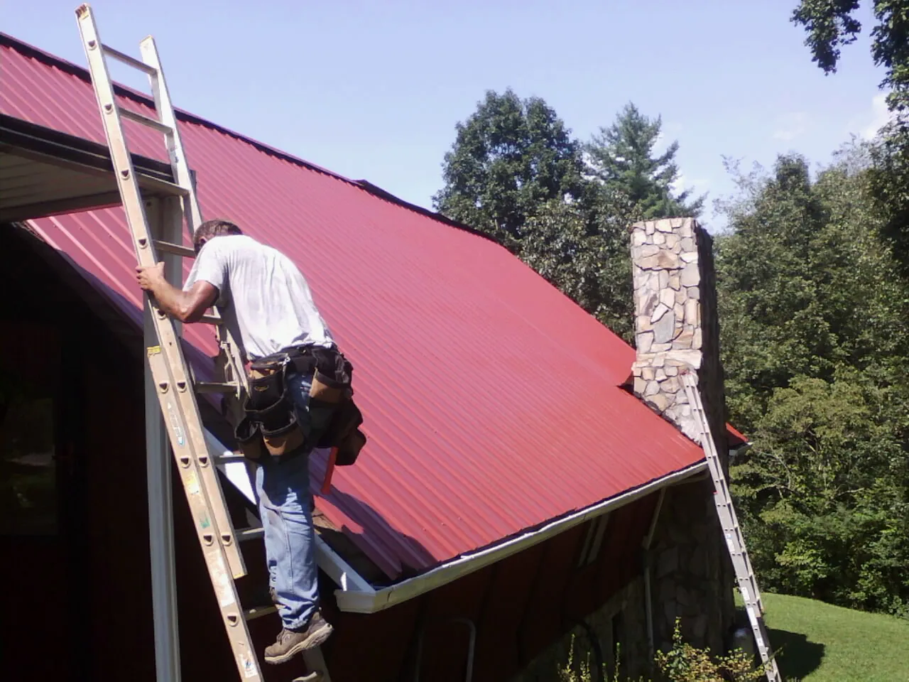 Roofer on a ladder beside a red metal roof and stone chimney — Soffit & Fascia Repair by Roofing Brothers in Franklinville